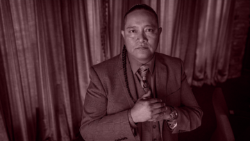 Portrait photograph of Lee Yaiva, an Indigenous man wearing a blue suit and sporting a braided ponytail; his hands are clasped together